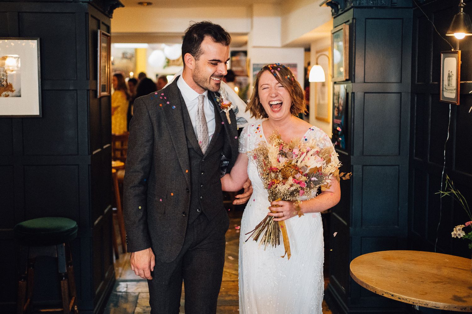 Joyful bride and groom laughing with confetti in a cozy pub venue, bride holding a dried floral bouquet.