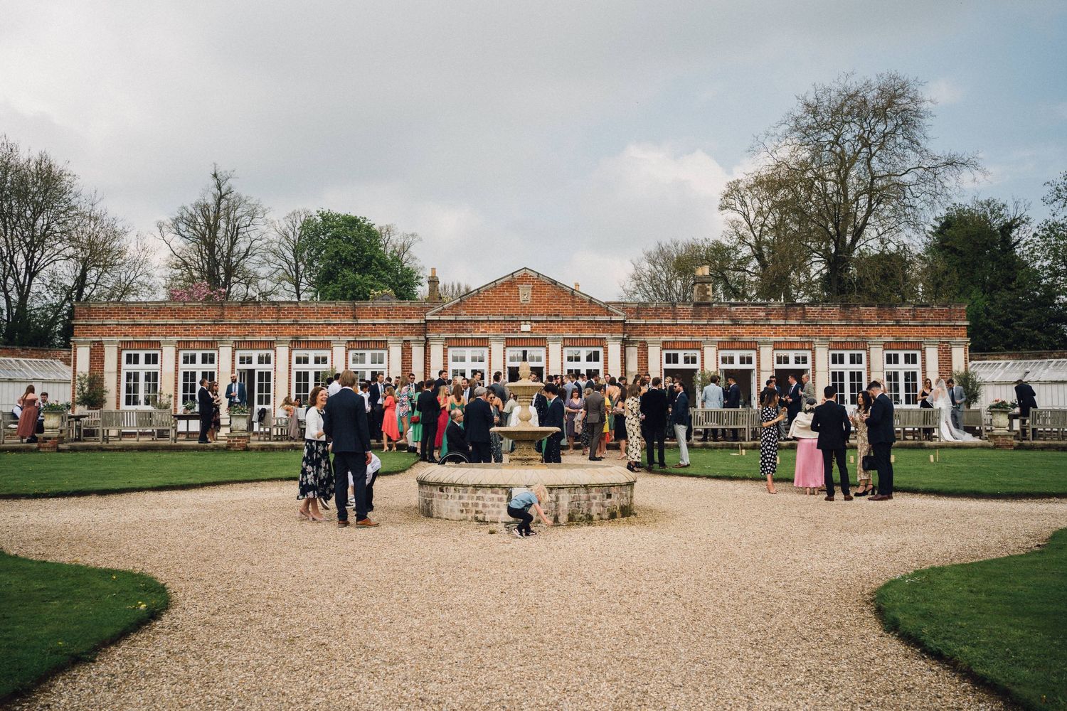Elegant Georgian orangery wedding venue with fountain courtyard, guests mingling outdoors amid manicured gardens and tall trees.