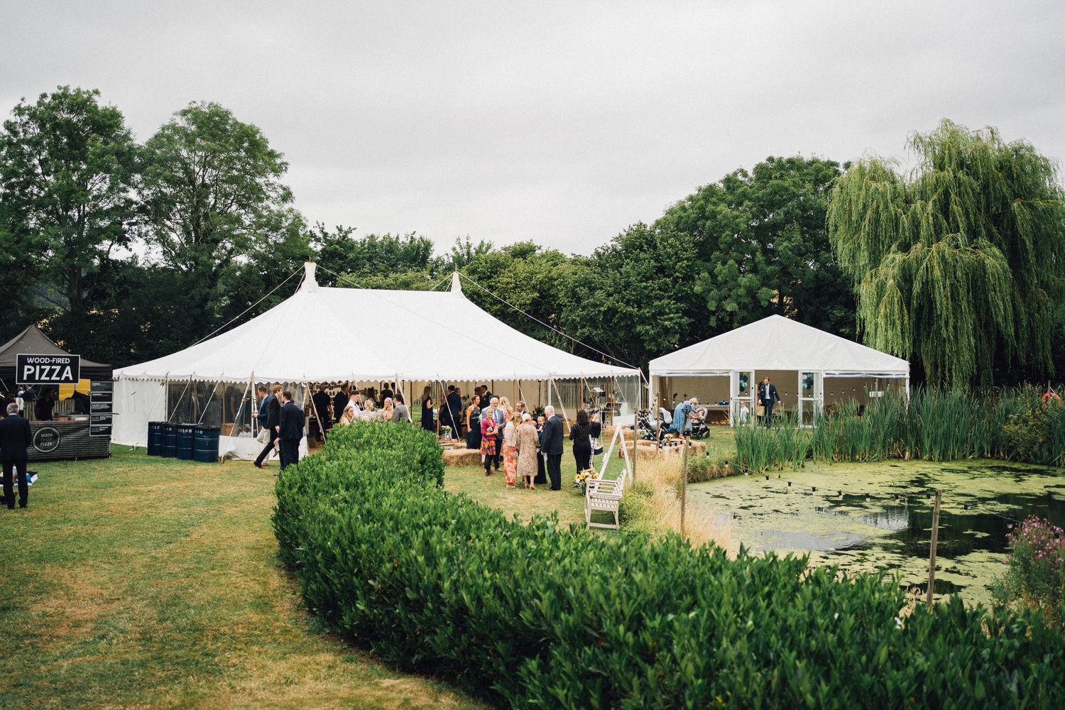 Elegant outdoor wedding marquee tent setup beside a tranquil lily pond, surrounded by lush greenery and willow trees.