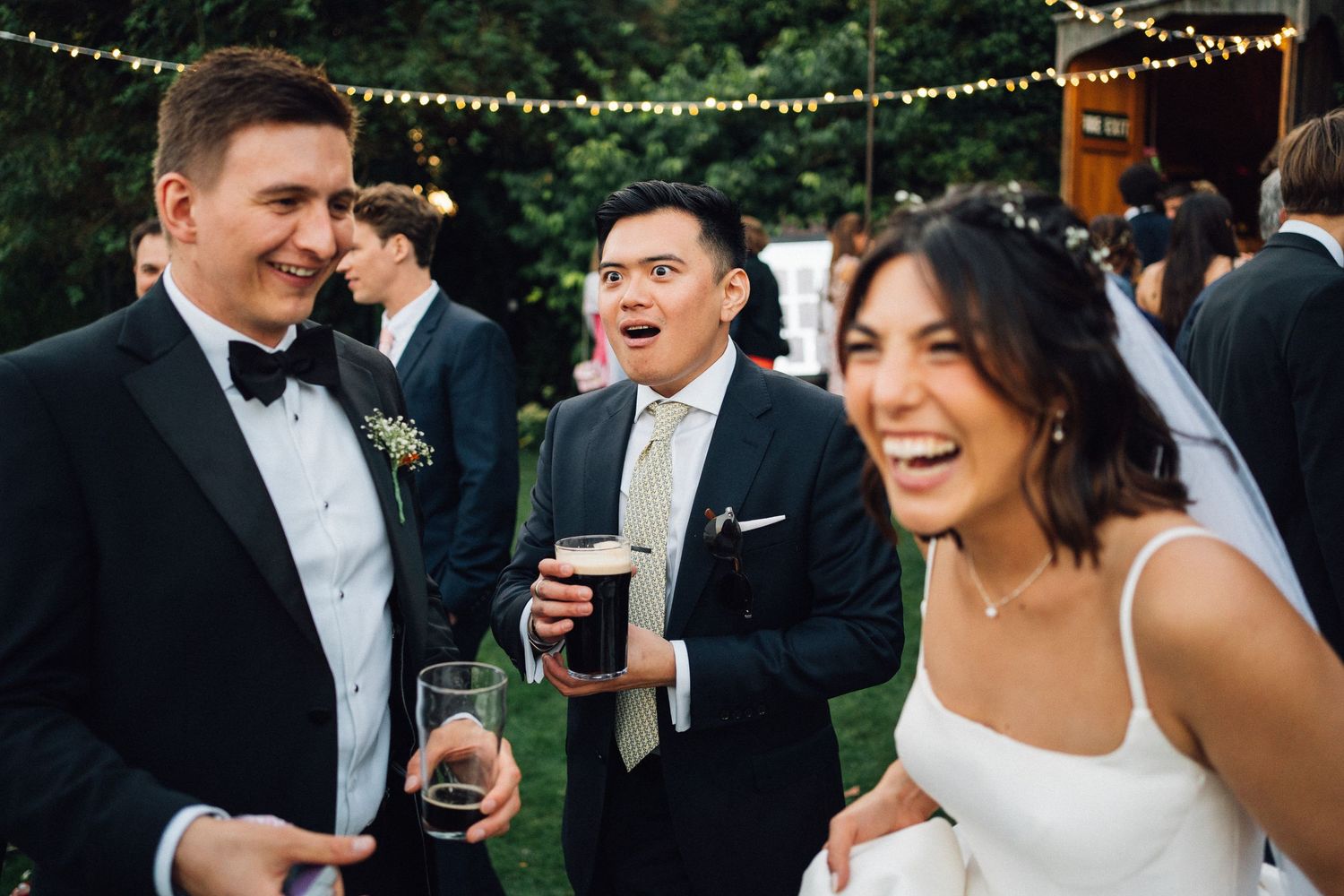 Wedding guests enjoy drinks and laughter at an outdoor reception under string lights during a festive celebration.