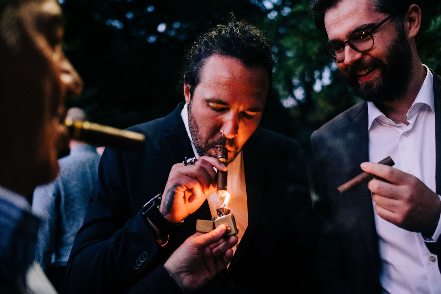 A group of friends gather outdoors to light cigars together in an evening setting.