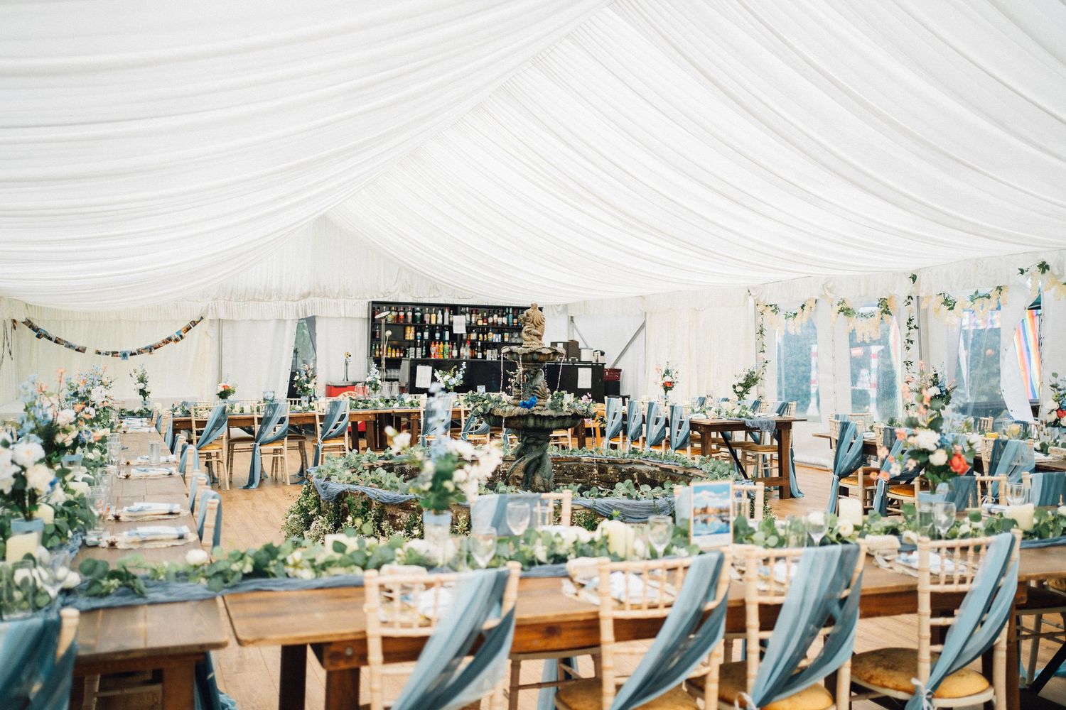 Wedding reception setup in an elegant white tent with rustic wooden tables, blue chairs and floral centerpieces.