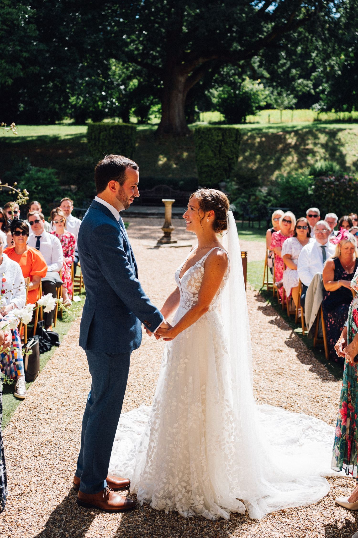 A wedding ceremony takes place outdoors on a gravel path surrounded by lush greenery and seated guests.