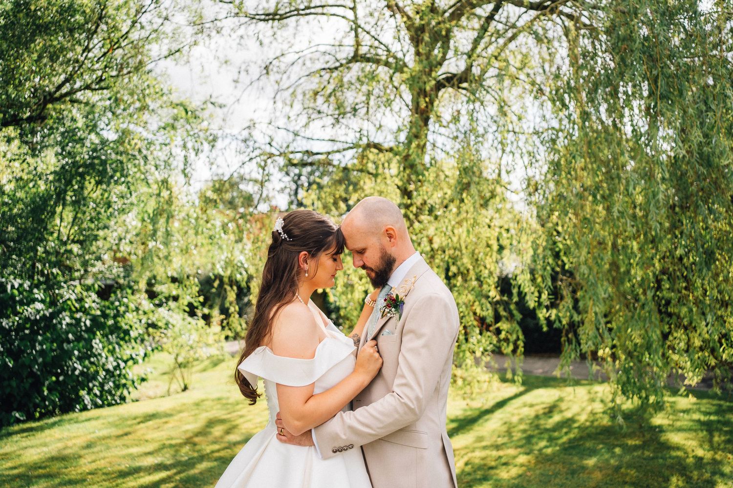 A romantic couple embraces under sunlit willow trees during a summer photo session in a garden setting.