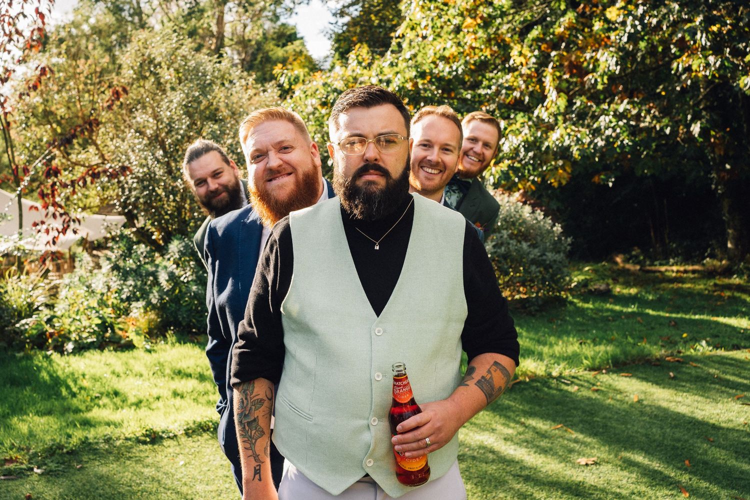 A group of groomsmen in casual attire share laughs and drinks while standing together in a sunny garden setting.