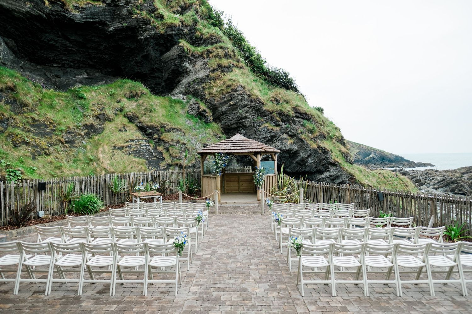 Outdoor wedding ceremony setup with white chairs facing an arch against dramatic coastal cliffs.