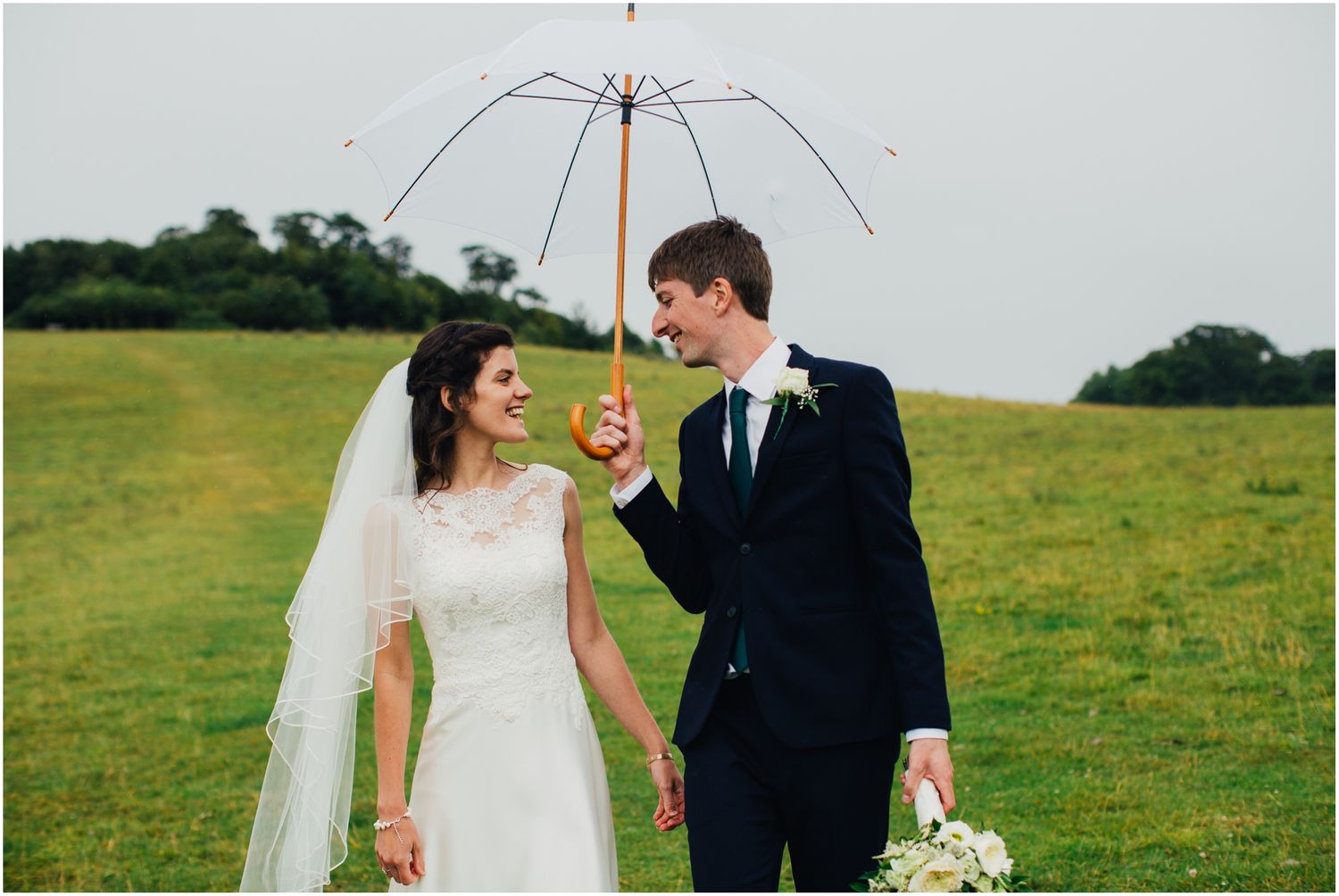 Couple holds clear umbrella while standing in green field during wedding photoshoot.