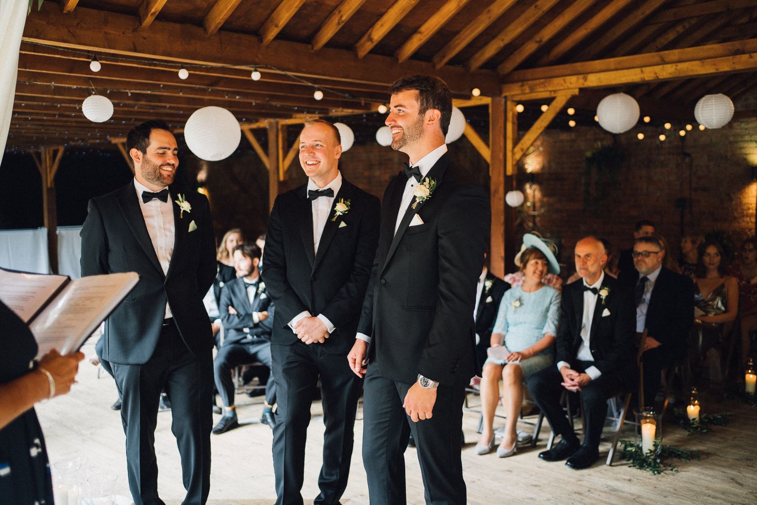 Wedding party poses in formal attire inside a rustic barn venue with decorative lighting.