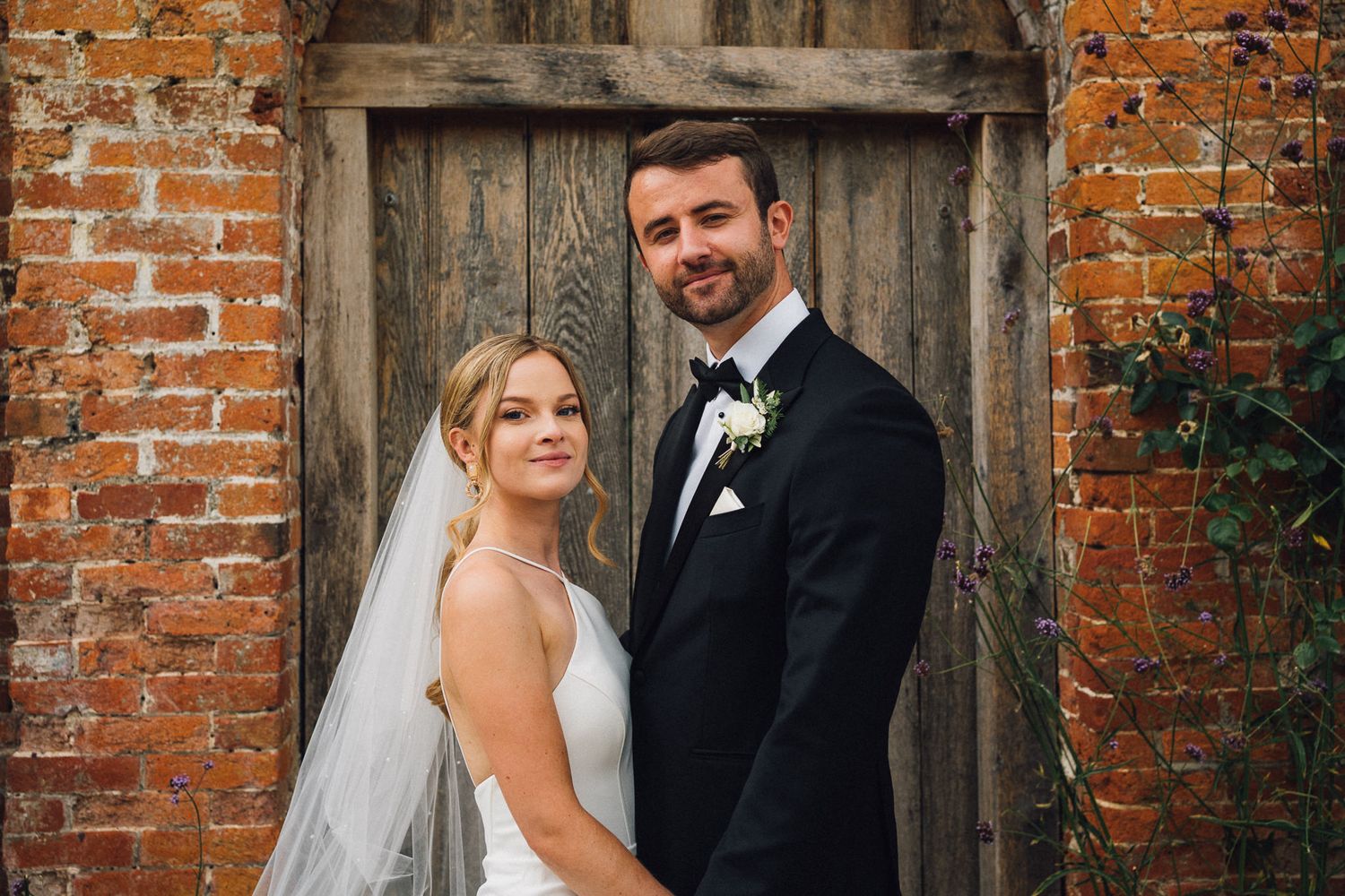 Wedding couple poses lovingly in front of an aged brick wall with wooden door.