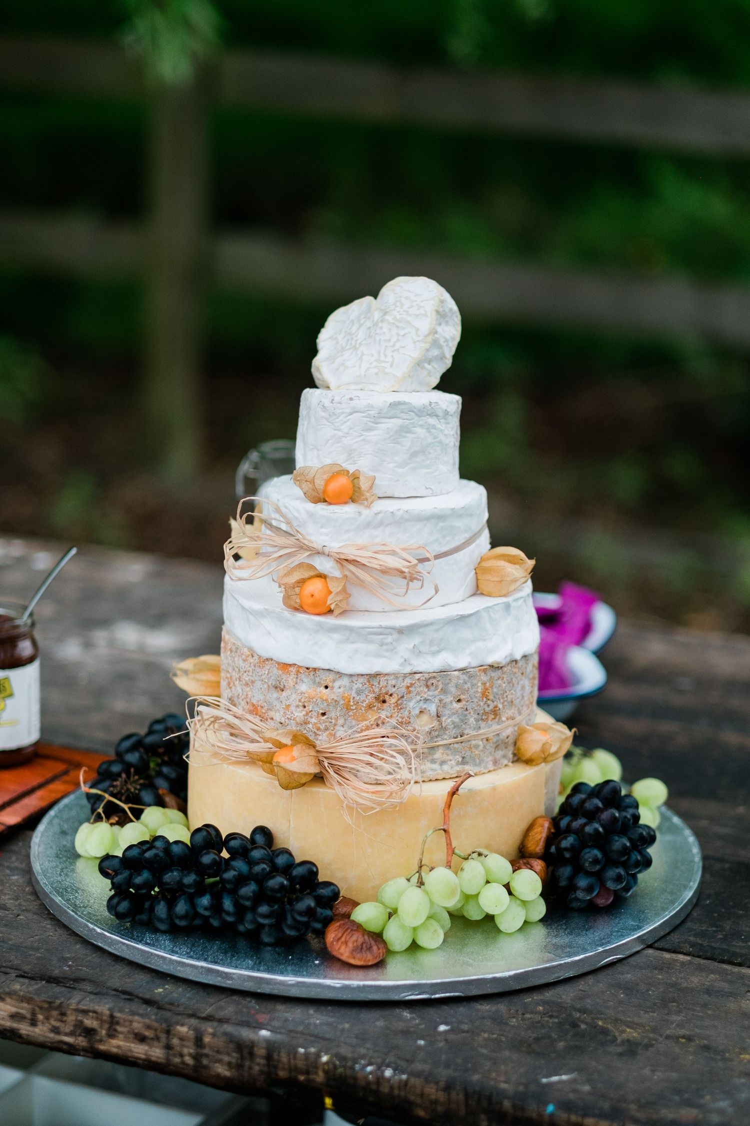 A tiered wedding cake decorated with lace, flowers and fresh grapes on a rustic outdoor table setting.