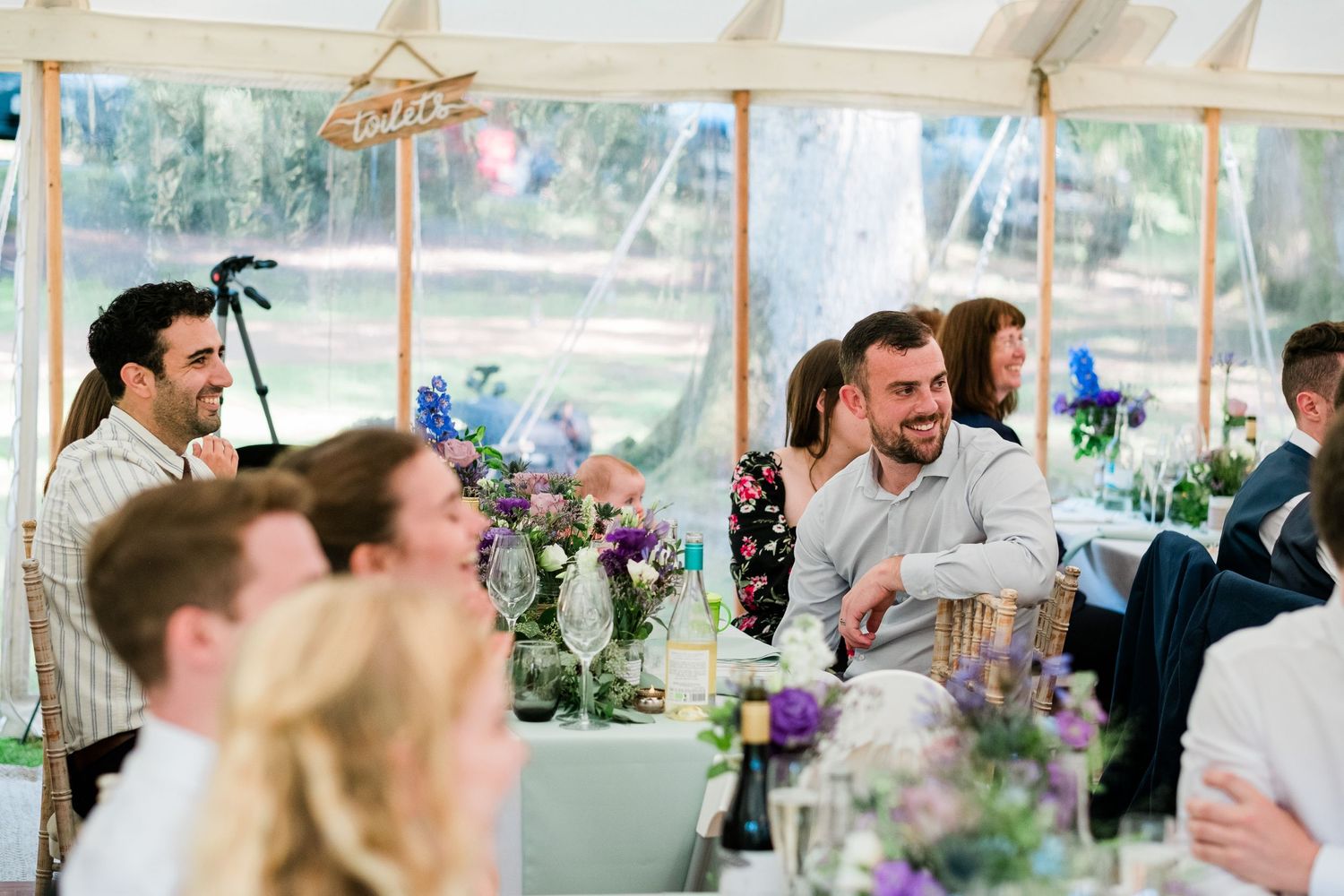 Wedding reception guests enjoying themselves under a white tent with string lights.