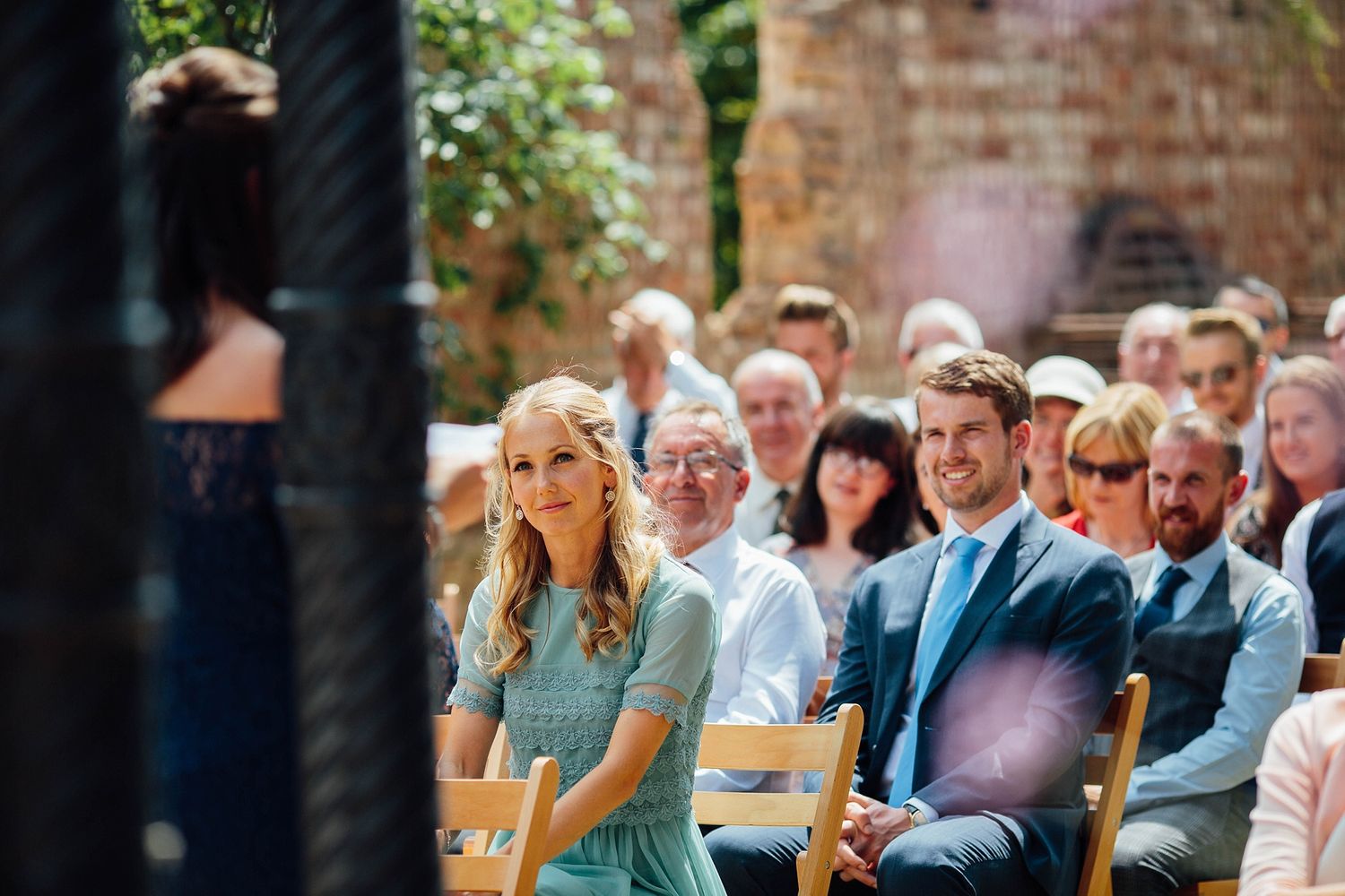 Emotional wedding guests react during an outdoor ceremony against a rustic brick wall backdrop.
