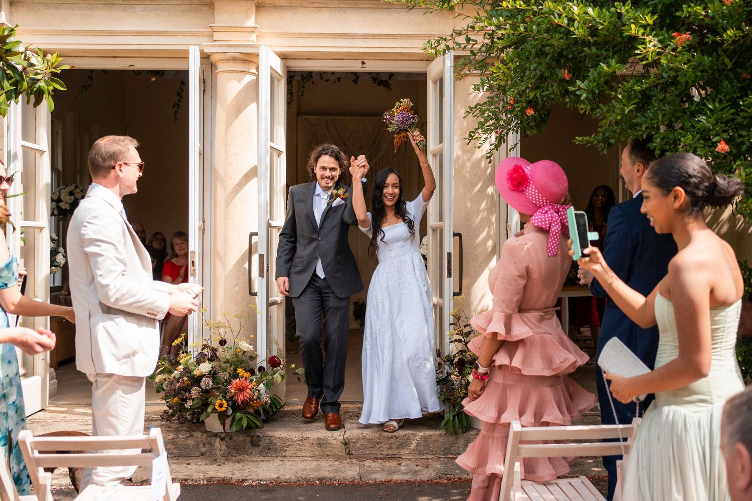 Joyful newlyweds exit wedding venue as guests celebrate, bride raising bouquet, woman in pink dress nearby.