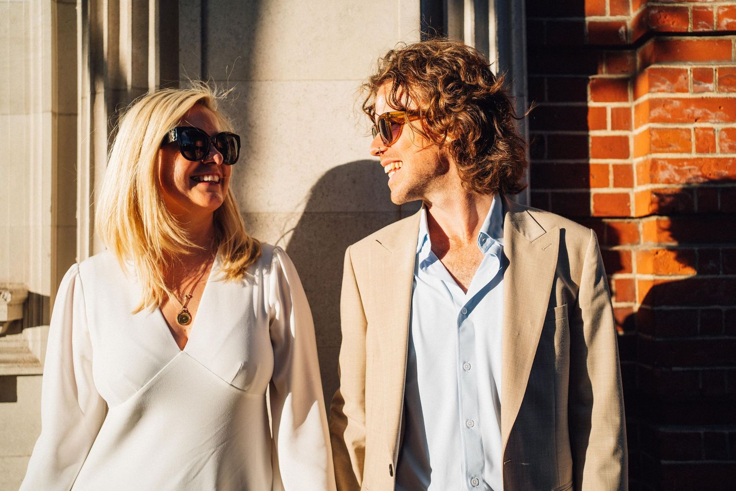 Two stylish young people wearing sunglasses and light-colored outfits smile at each other against a brick wall.