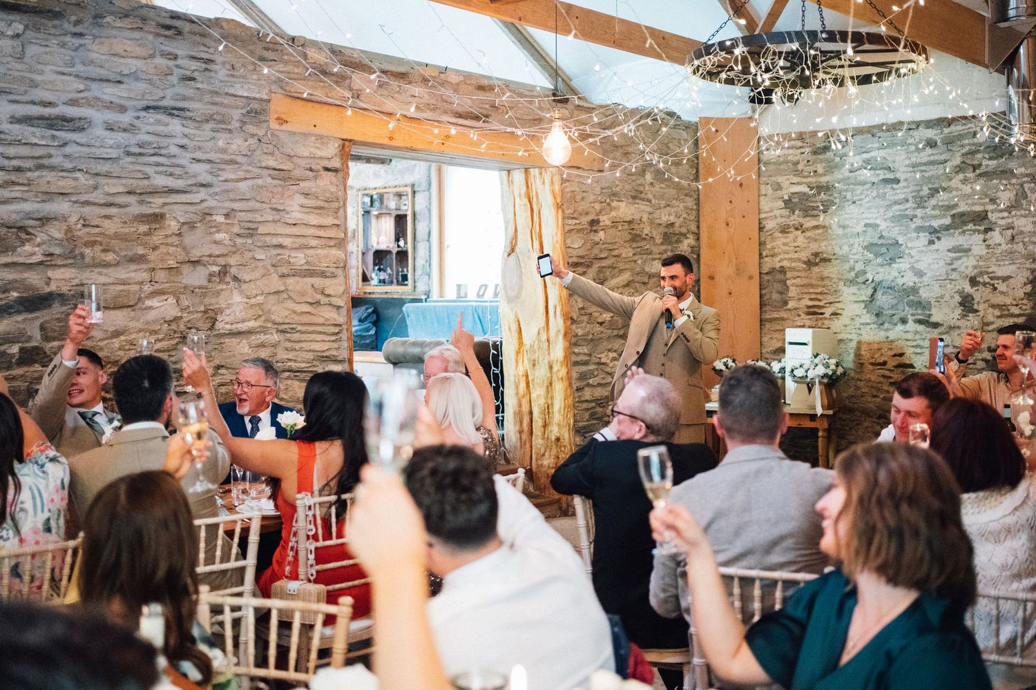 Wedding guests celebrate and applaud during a reception in a rustic stone-walled venue with wooden beams.