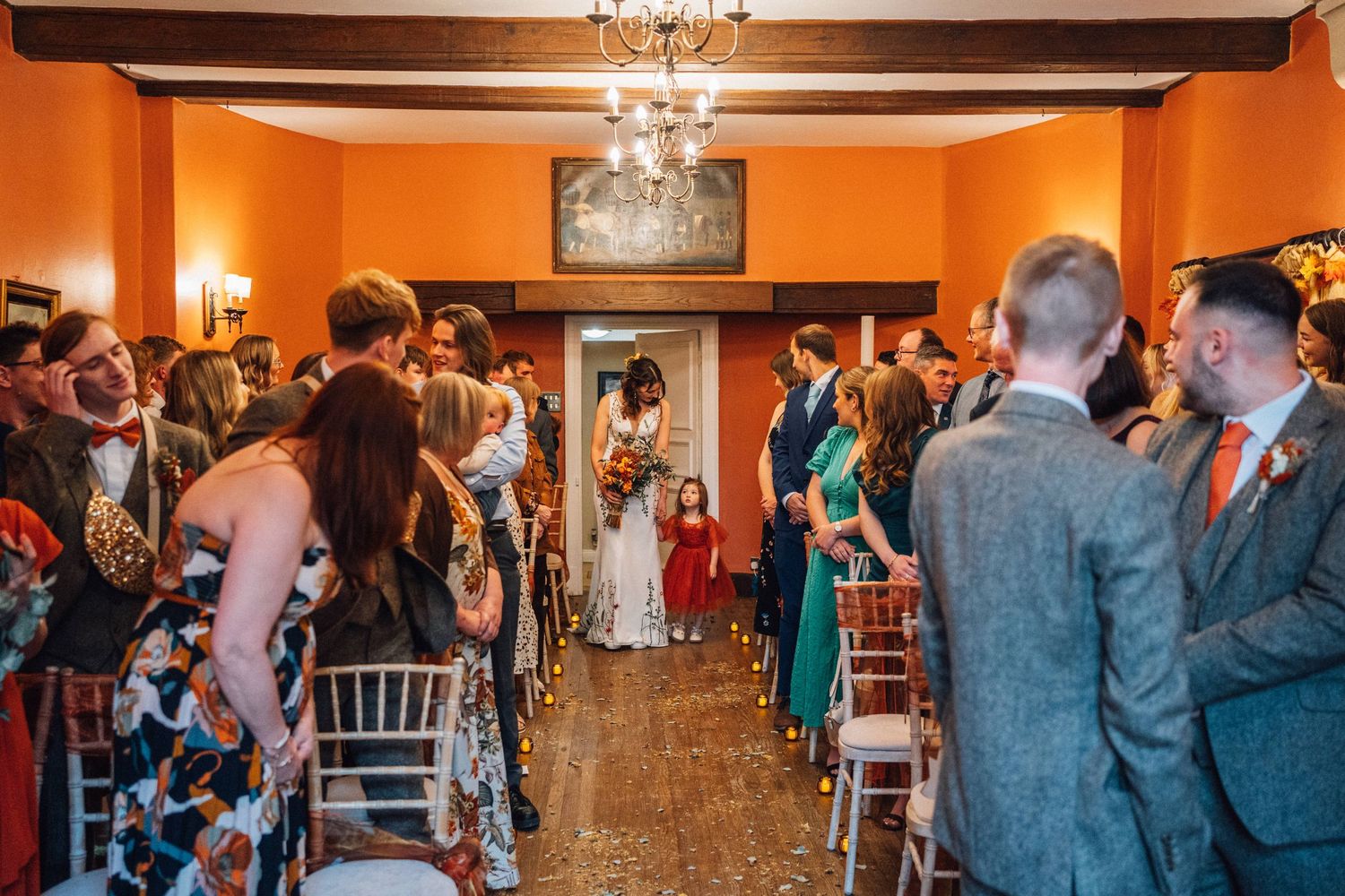 Wedding ceremony guests gather in an orange-walled room with chandelier and white chairs for an indoor celebration.