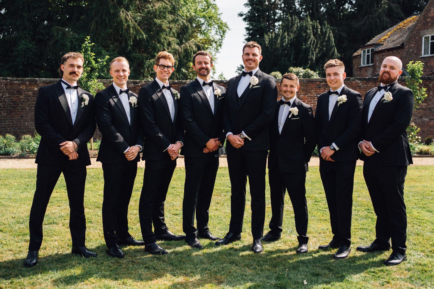 Groomsmen in black tuxedos and bow ties pose together on a sunny lawn for a formal wedding photo.