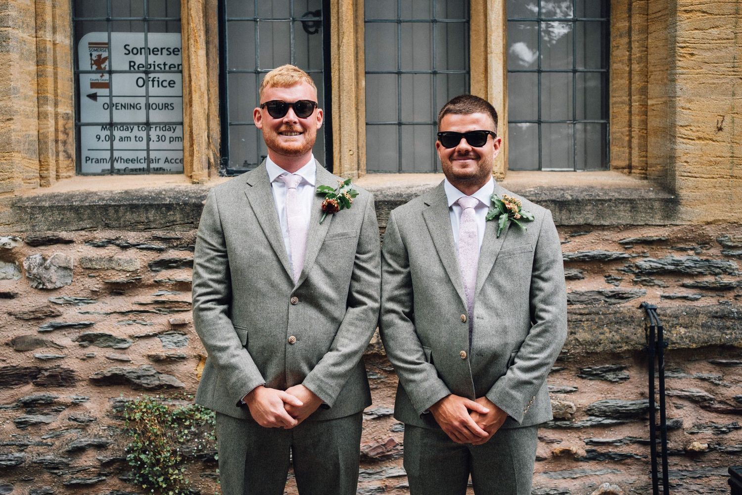 Two people in gray suits and sunglasses stand together smiling in front of a stone wall.