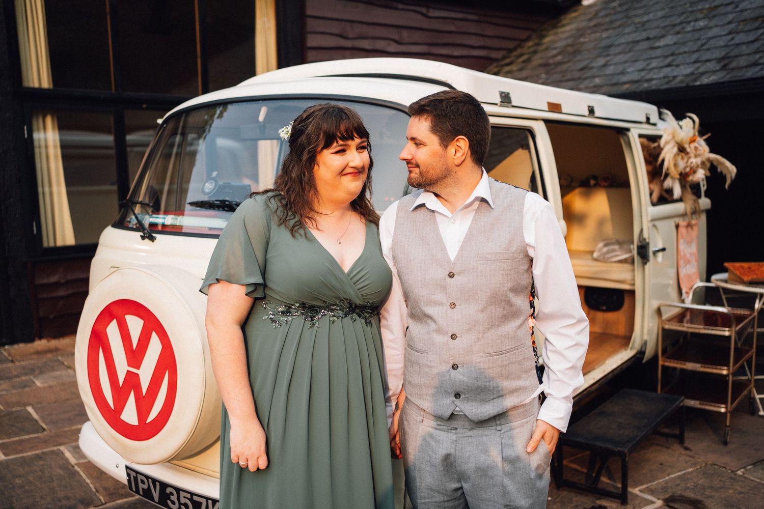 Couple in coordinated sage green and gray outfits posing together in front of vintage camper trailer.