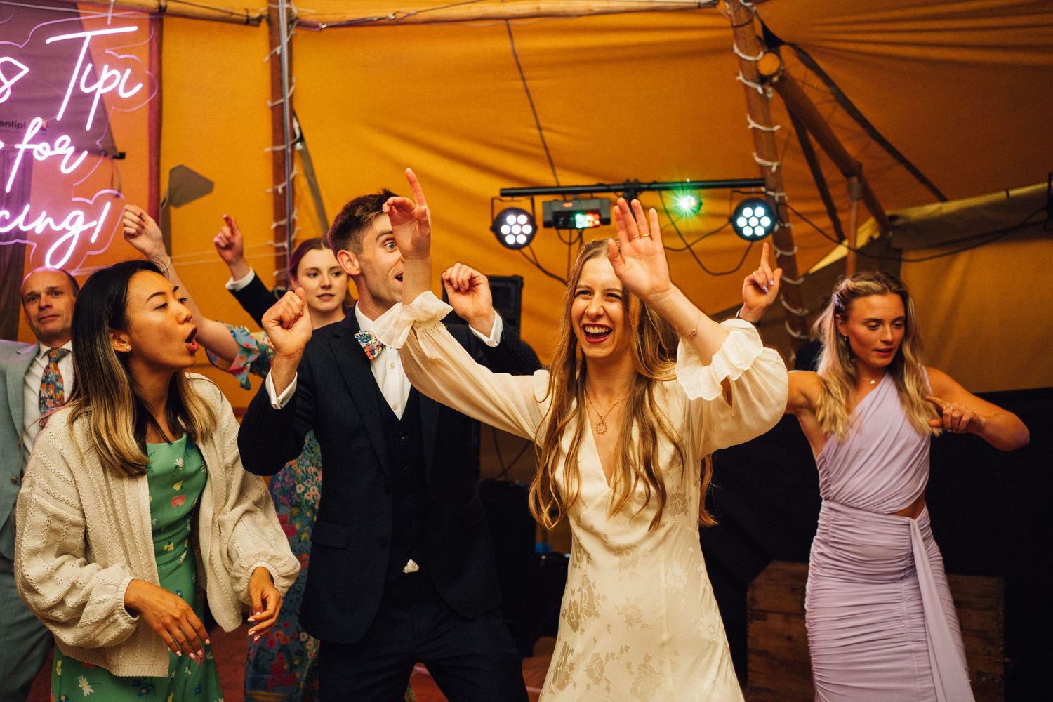 Wedding party dancing and singing enthusiastically under tent with neon sign.