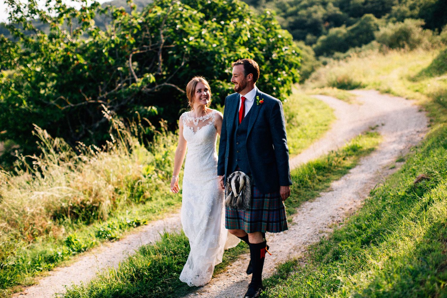 A couple walks down a scenic country path surrounded by lush greenery and rolling hills.