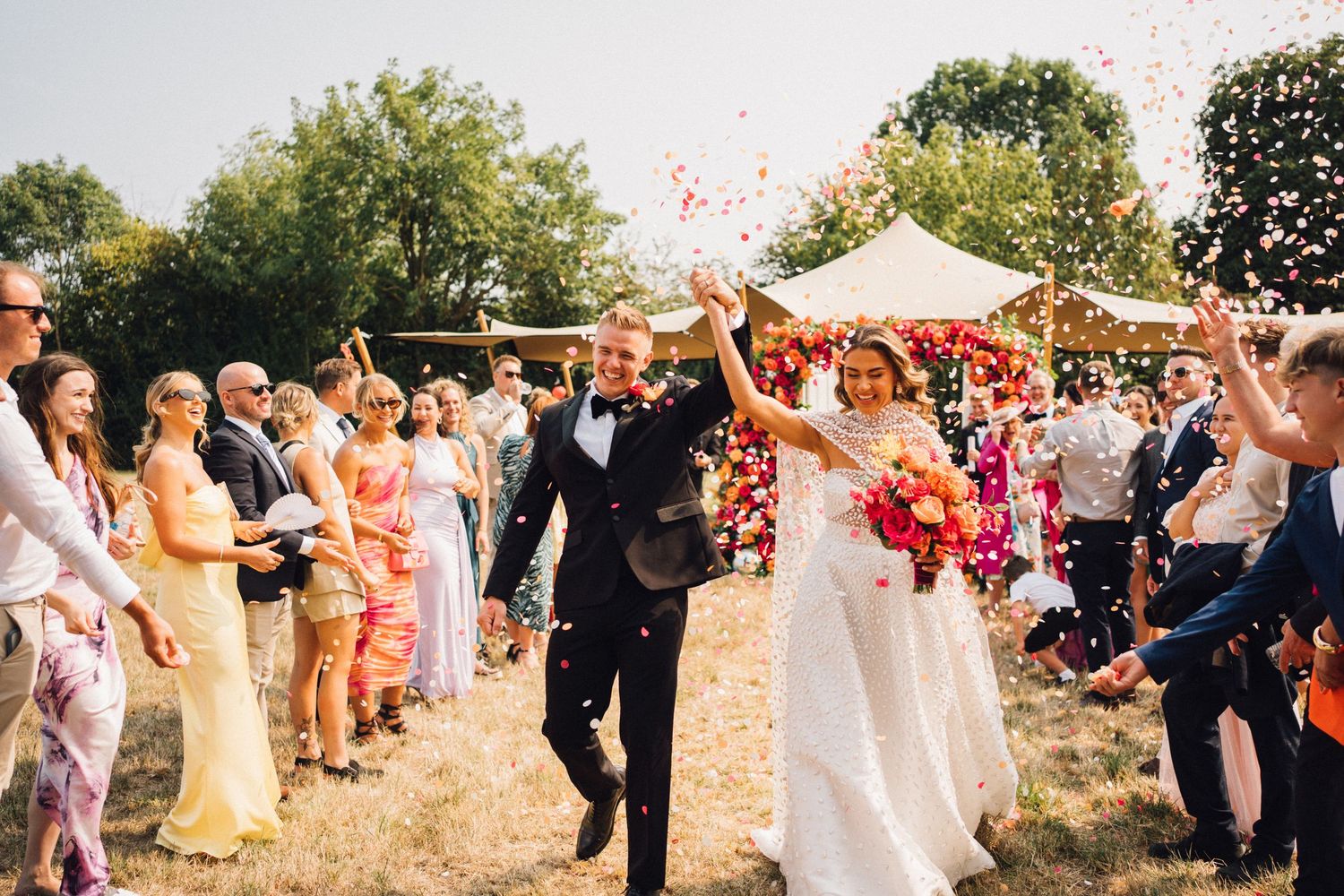 Newlyweds celebrate with confetti and joy as they walk down an outdoor aisle lined with guests at a summer wedding.