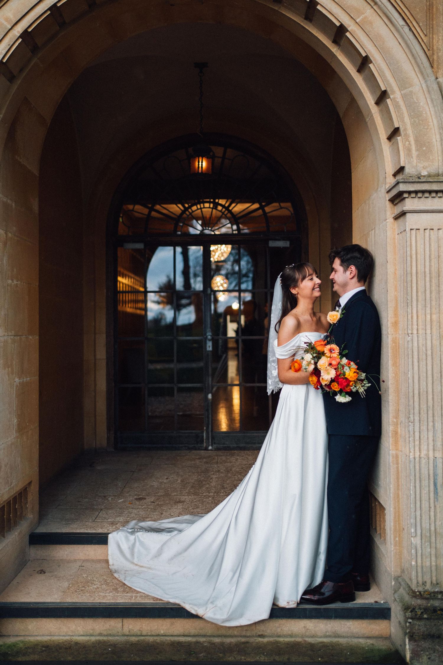Bride and groom share a romantic moment under a stone arch doorway, bride holding vibrant orange and red bouquet.