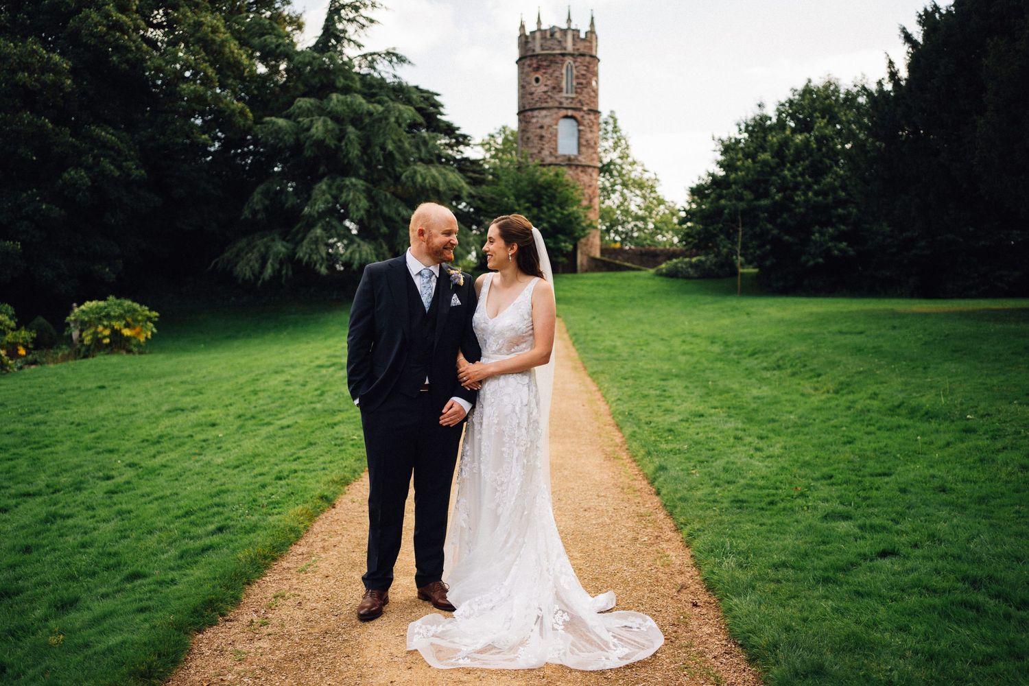 Bride and groom holding hands on a garden path with a historic stone church tower in the background.