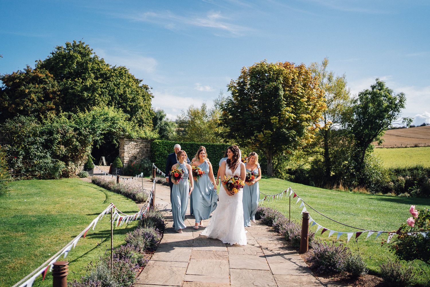 Bride and bridesmaids in blue dresses walking along a garden path at a sunny outdoor wedding venue.