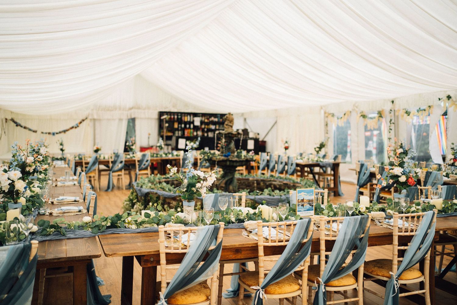 Long banquet tables with blue chairs set up under white tent ceiling for an elegant wedding reception.