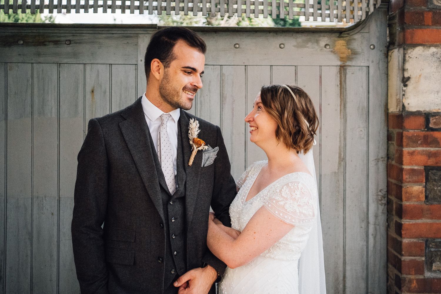 A joyful wedding couple shares a candid moment together against a rustic gray door and brick wall backdrop.