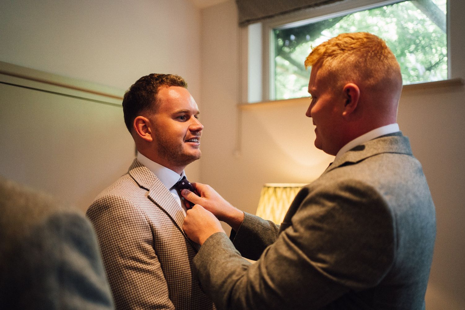 Two people in gray suits share a tender moment as one adjusts the other's tie before a formal event.