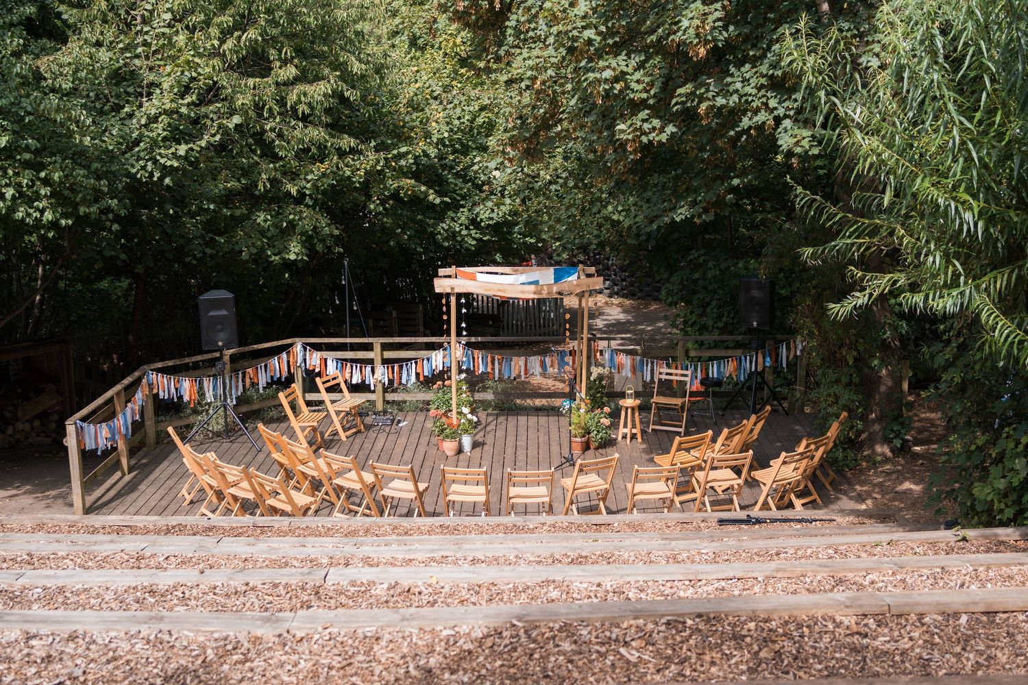 Wooden deck with chairs arranged in a circle, decorated with colorful bunting, surrounded by lush green trees.