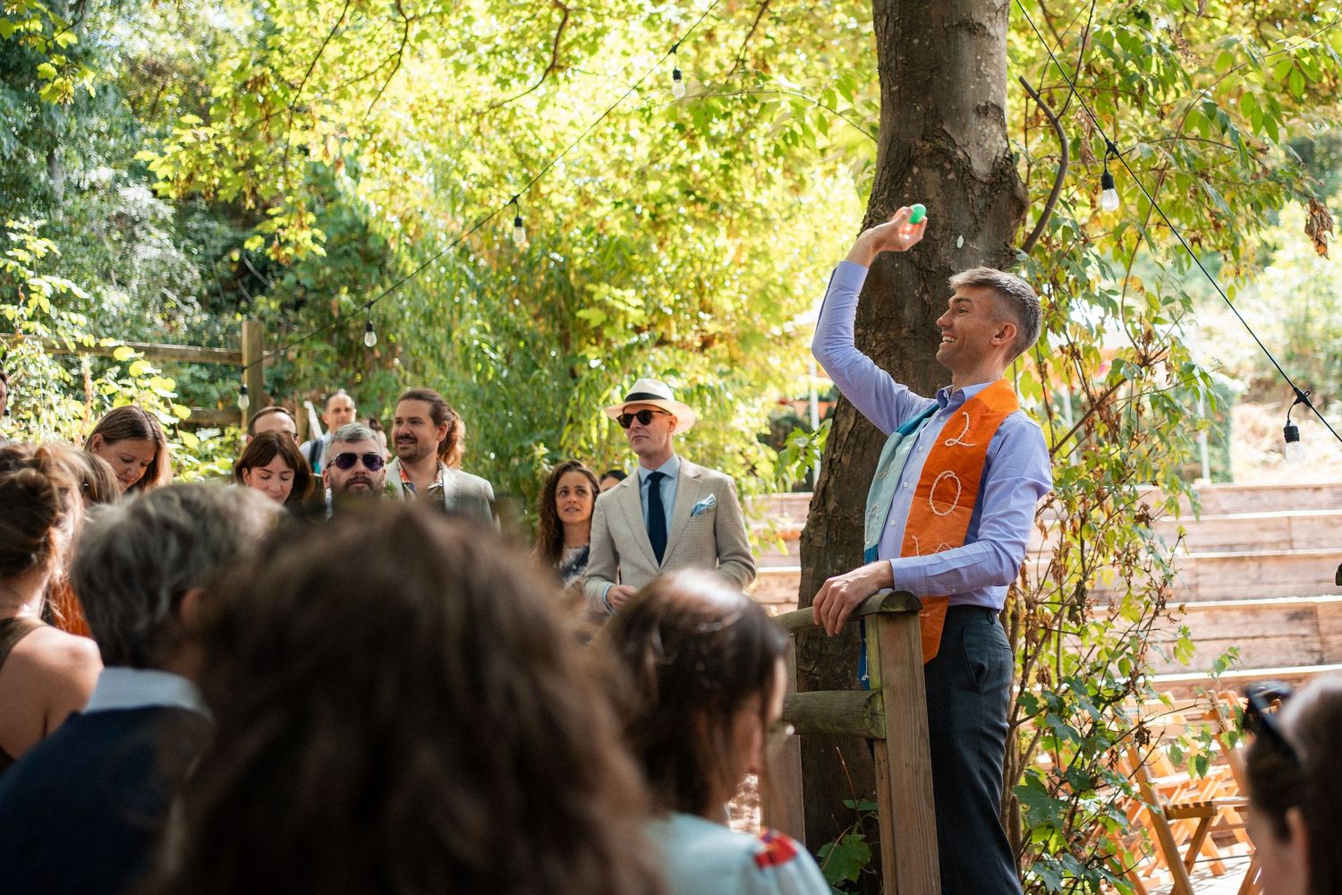 Man in orange sash raises a drink at an outdoor wedding ceremony surrounded by guests in a lush garden setting.