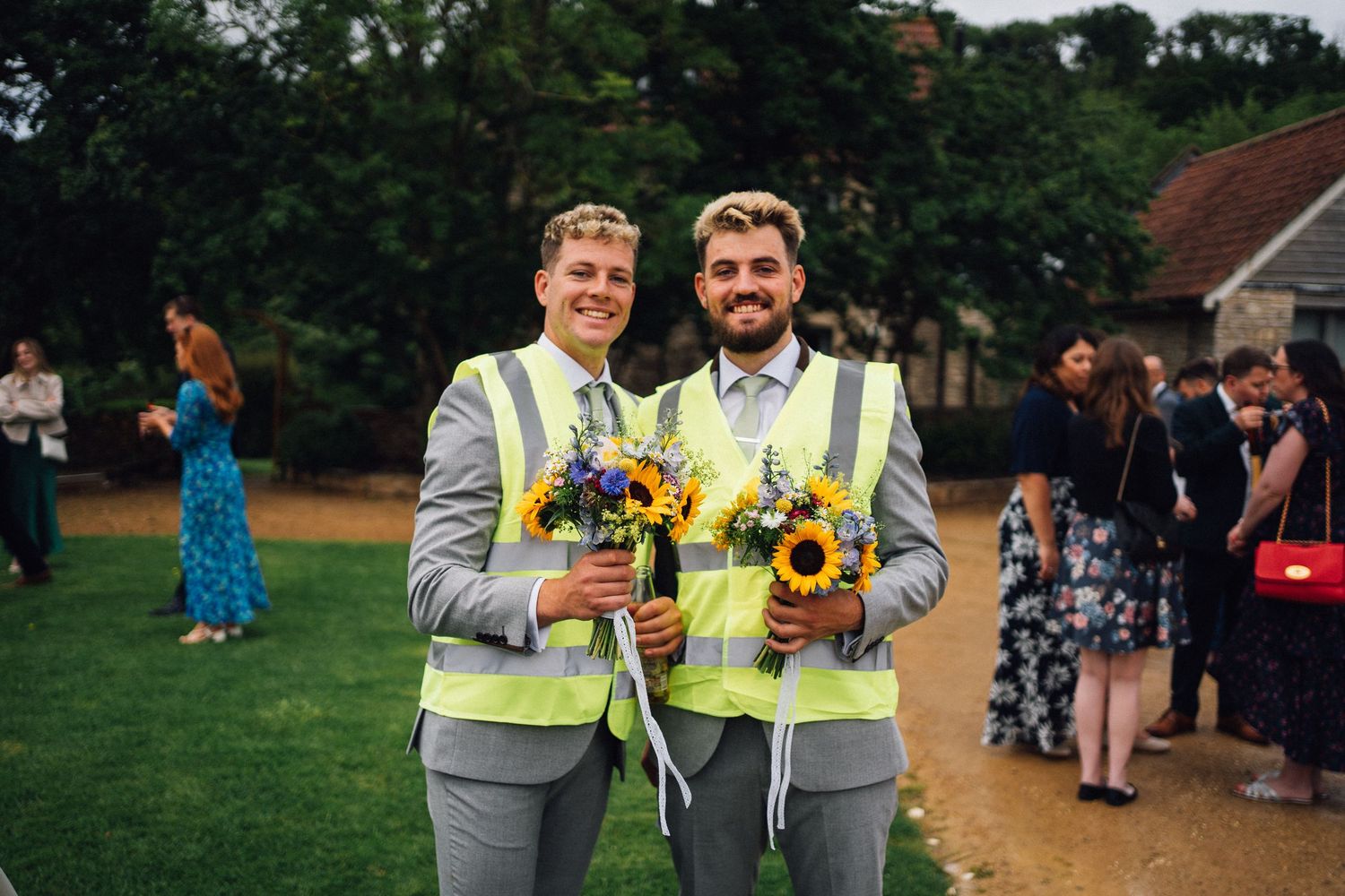 Two people in matching gray suits hold sunflowers at outdoor wedding celebration.