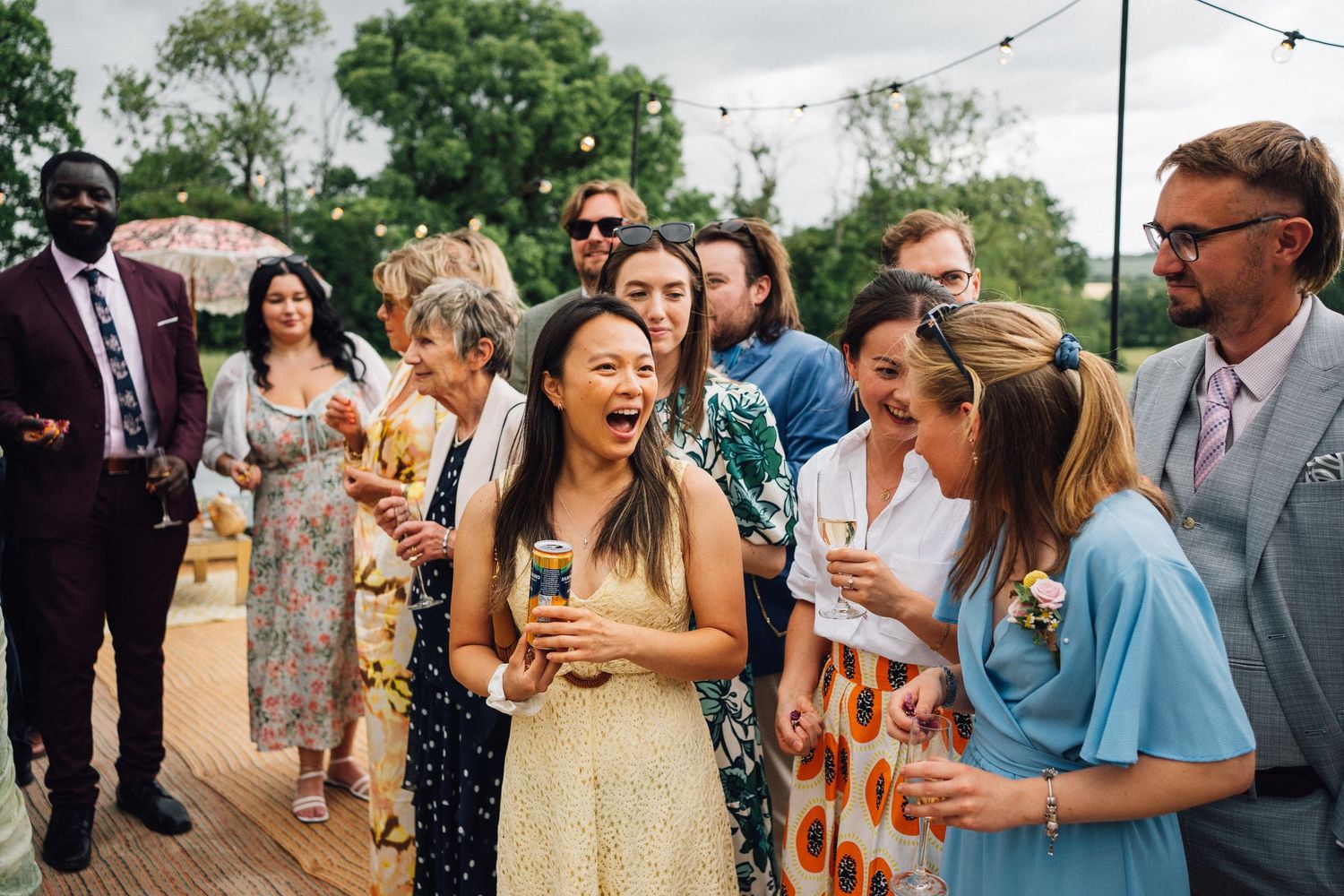 Wedding guests laughing and socializing on a wooden deck during an outdoor reception.