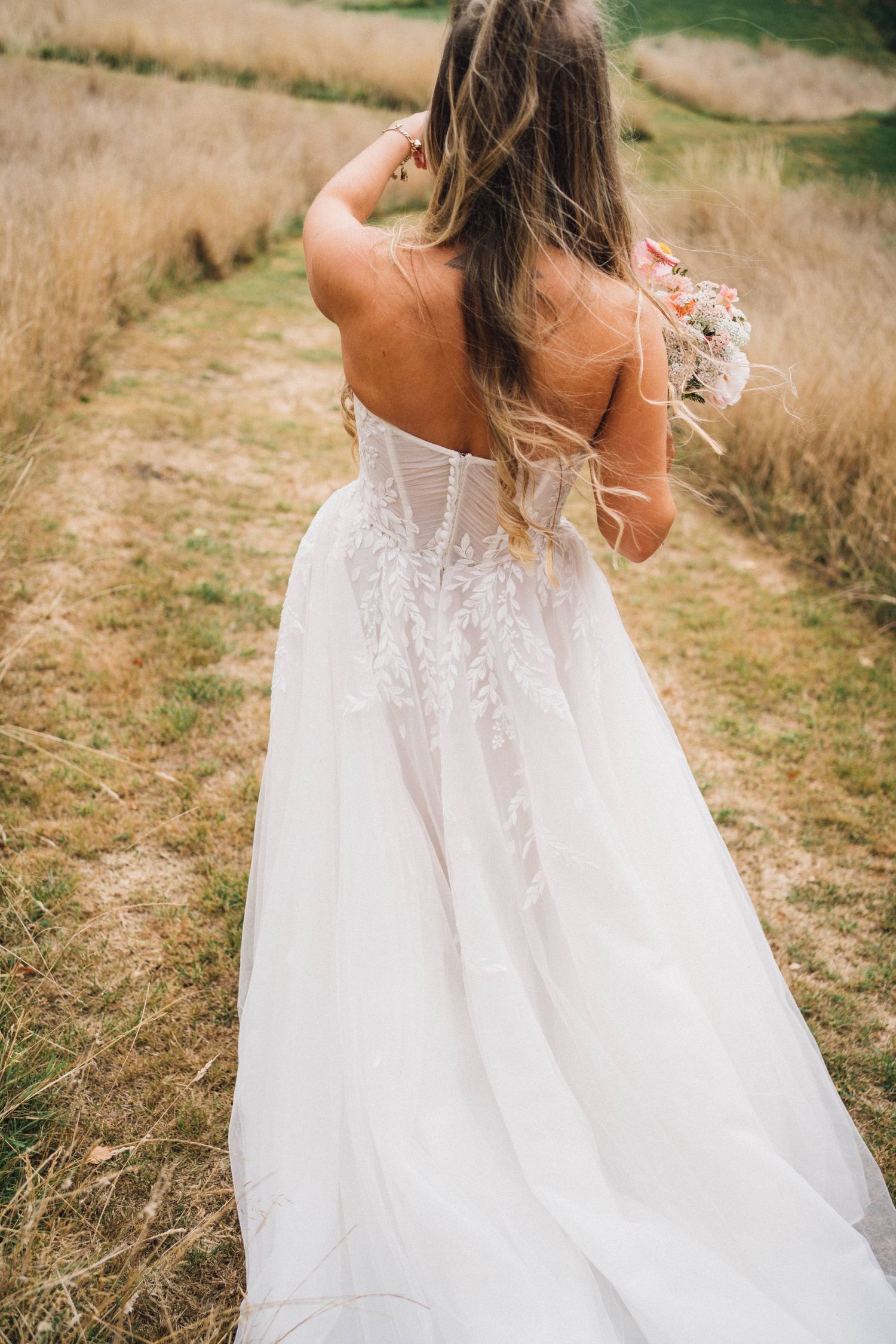 Back view of a bride in a detailed white wedding gown with lace details standing in a golden field.