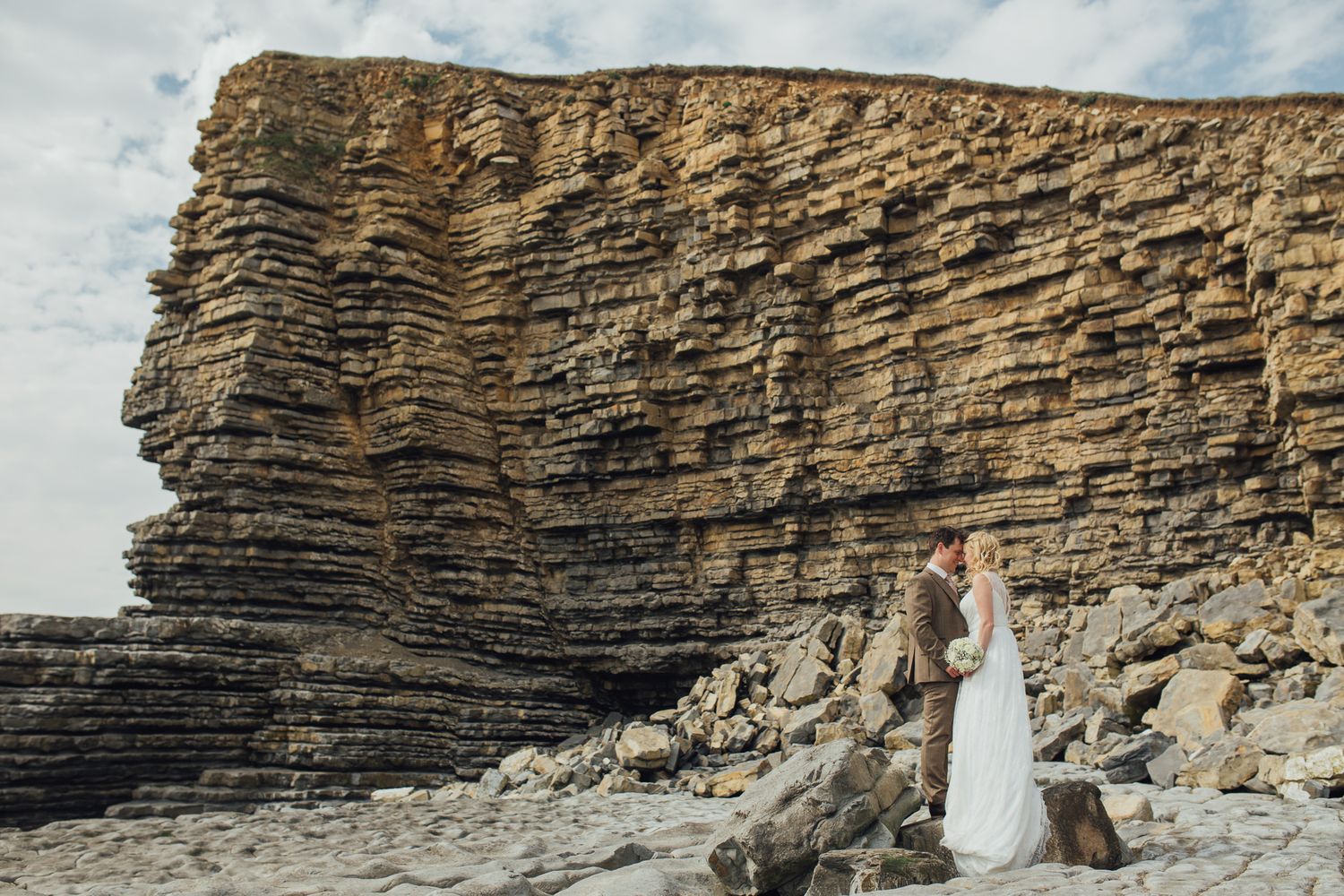 Wedding couple poses against dramatic layered rock formations at coastal cliffs.