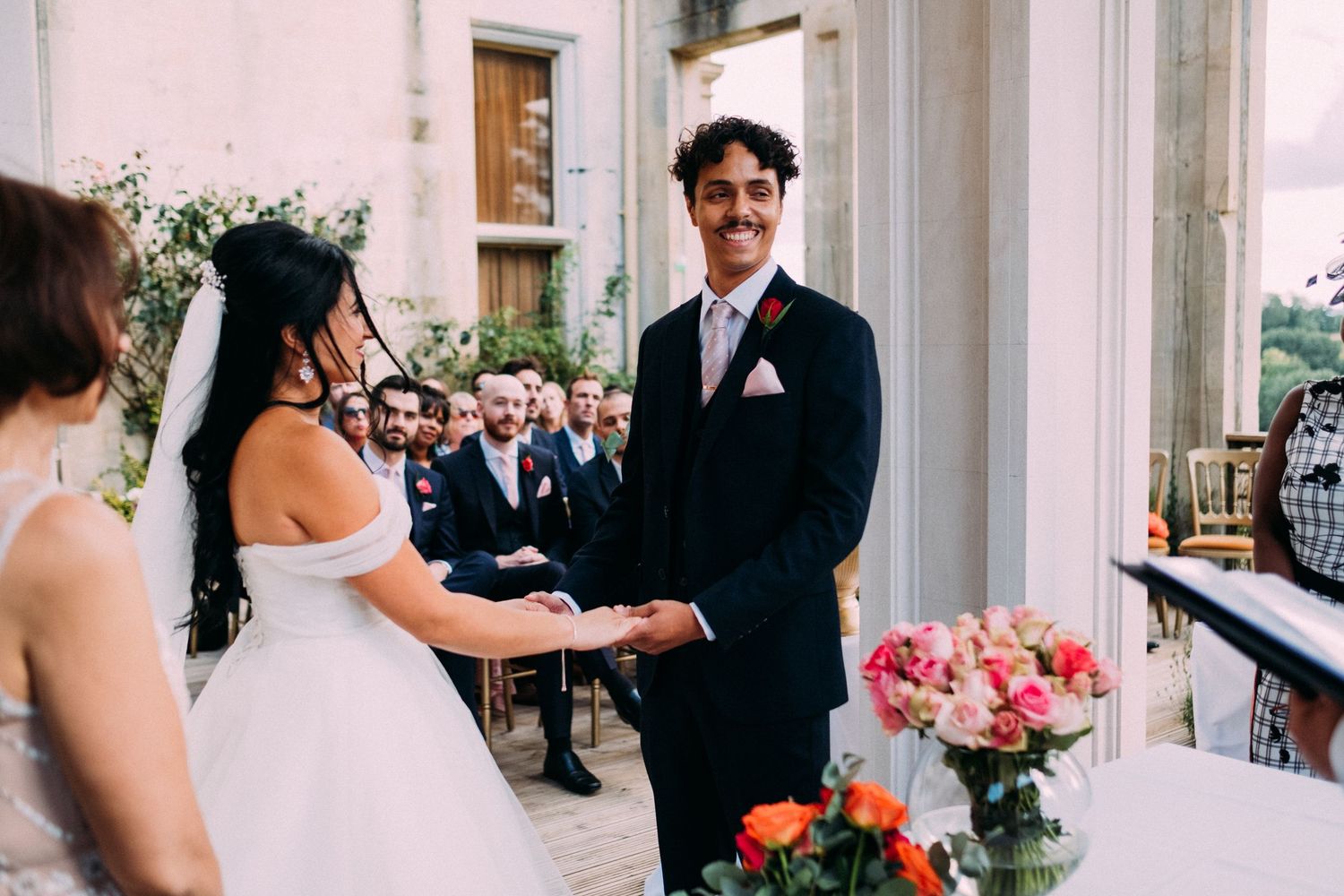 An intimate outdoor wedding ceremony with pink floral decorations and white architectural columns.