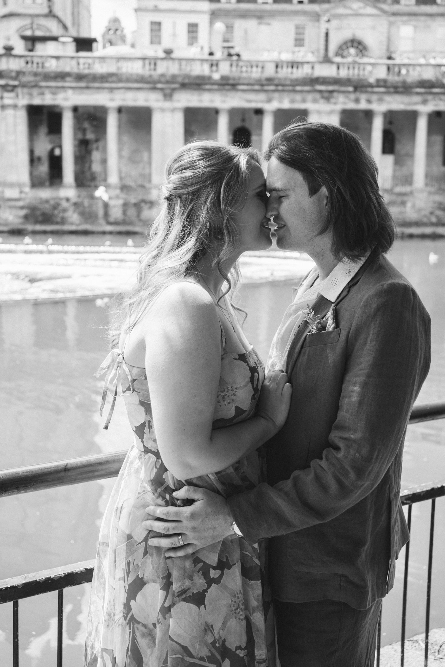 A romantic couple shares an intimate moment along a waterfront railing in black and white.