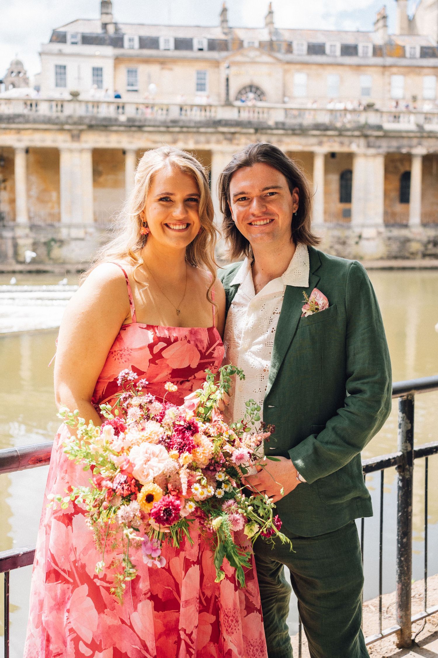 Couple poses by historic stone balustrade overlooking water with vibrant pink floral bouquet.