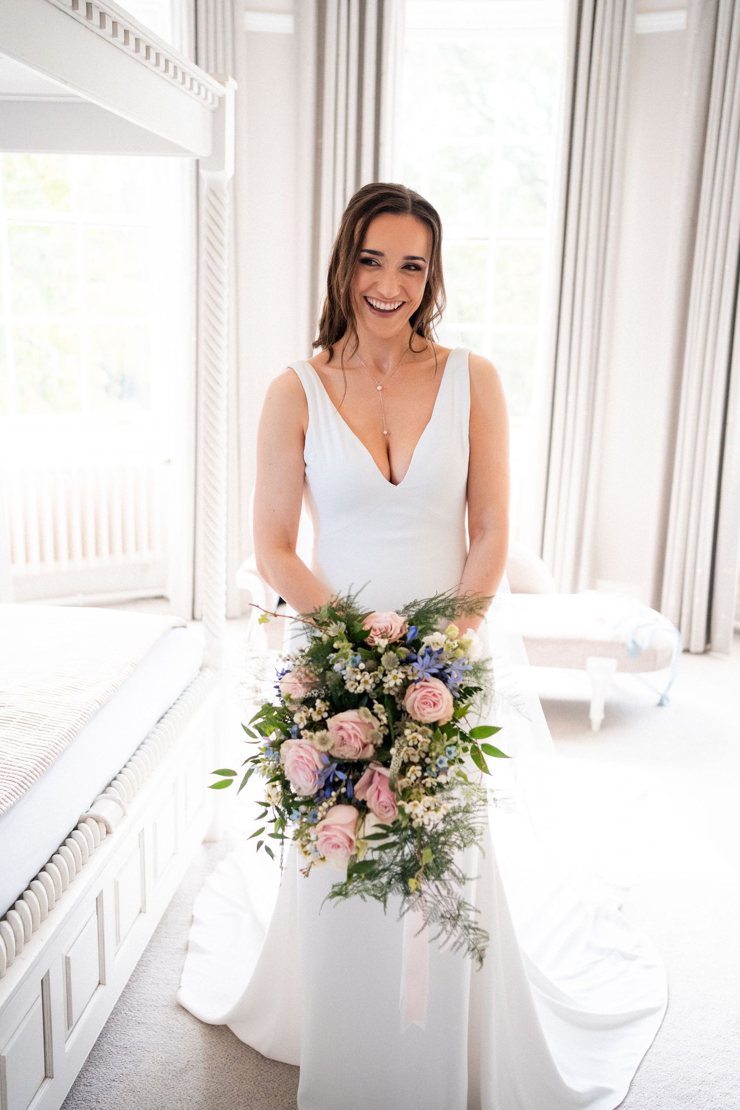 Smiling bride in white gown holding cascading pink and blue floral bouquet standing in bright elegant room.