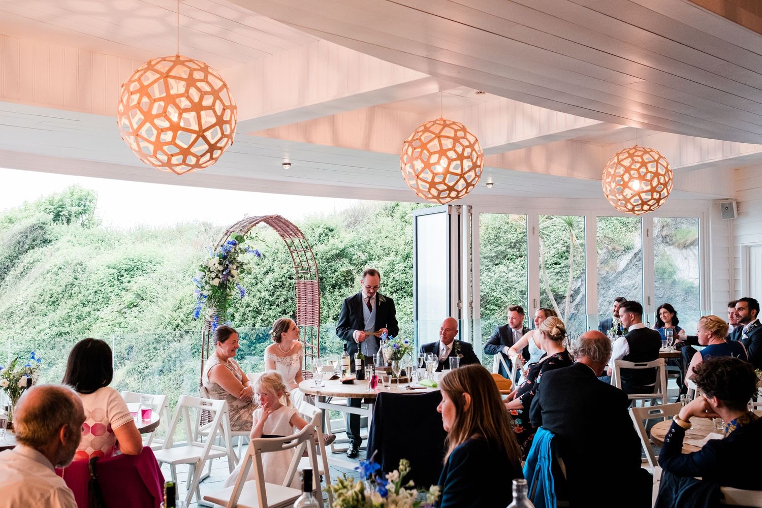 Wedding reception space decorated with geometric orange pendant lights and large windows.