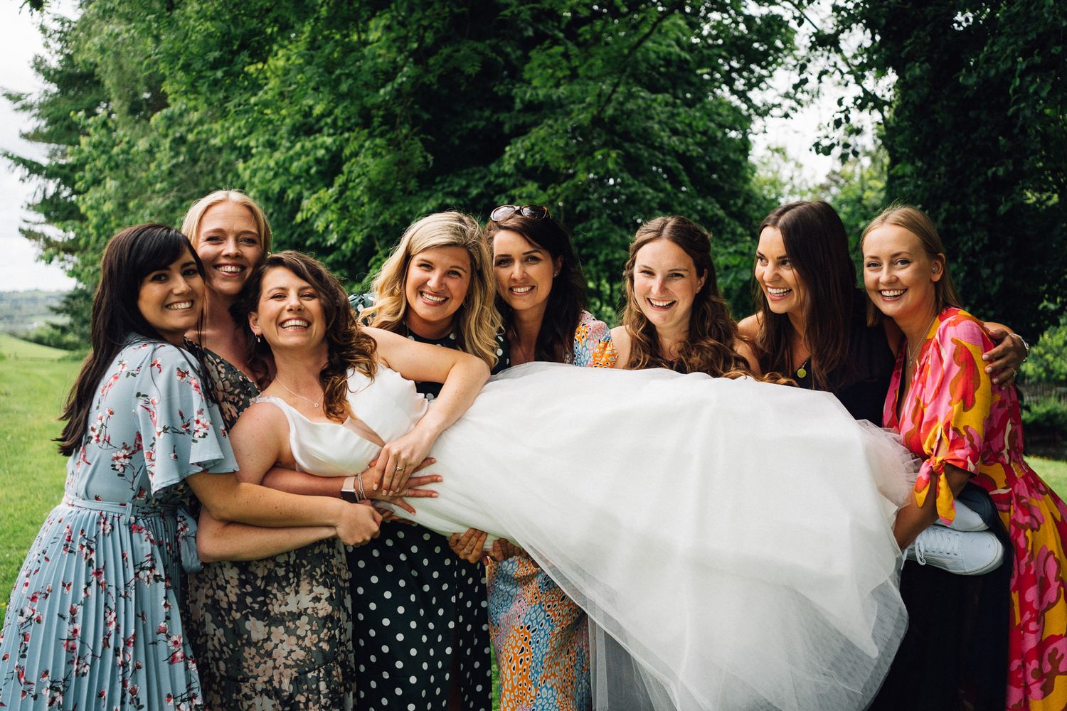Bridesmaids in colorful dresses holding up the bride's white wedding dress train outdoors.