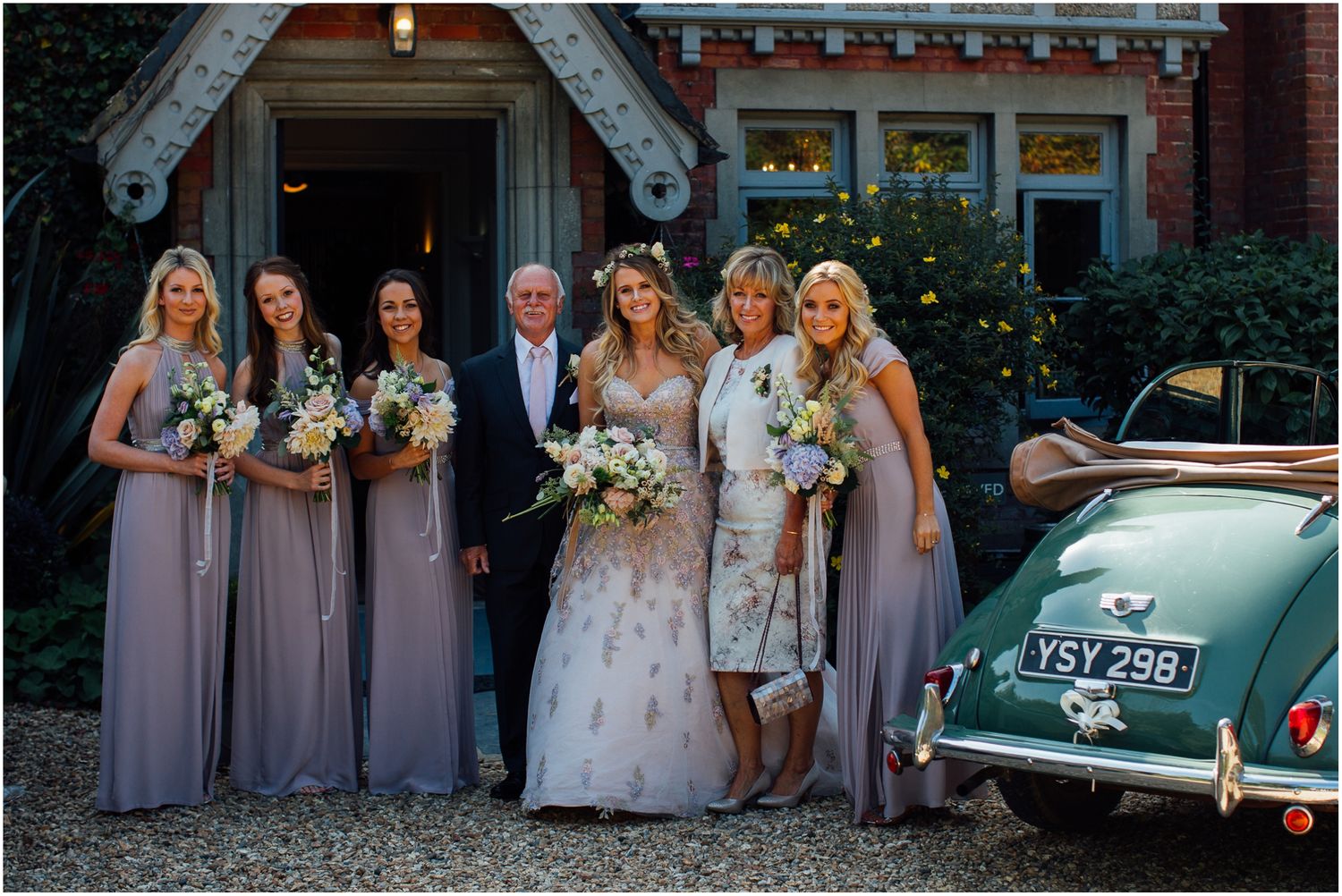 Wedding party poses with vintage green car outside a brick building.