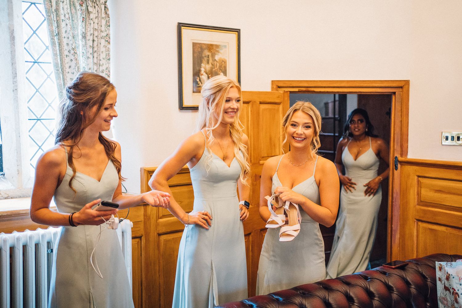 Bridesmaids in silver dresses gather in a room with wooden furniture and natural light.