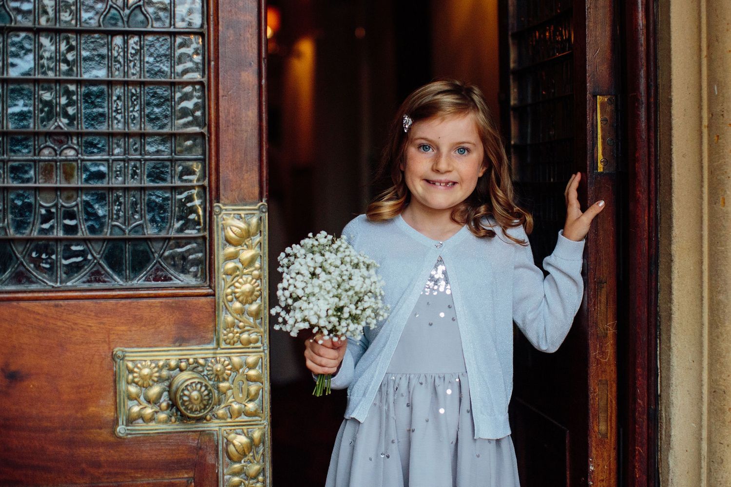 Young flower girl in light blue dress standing by wooden doors.