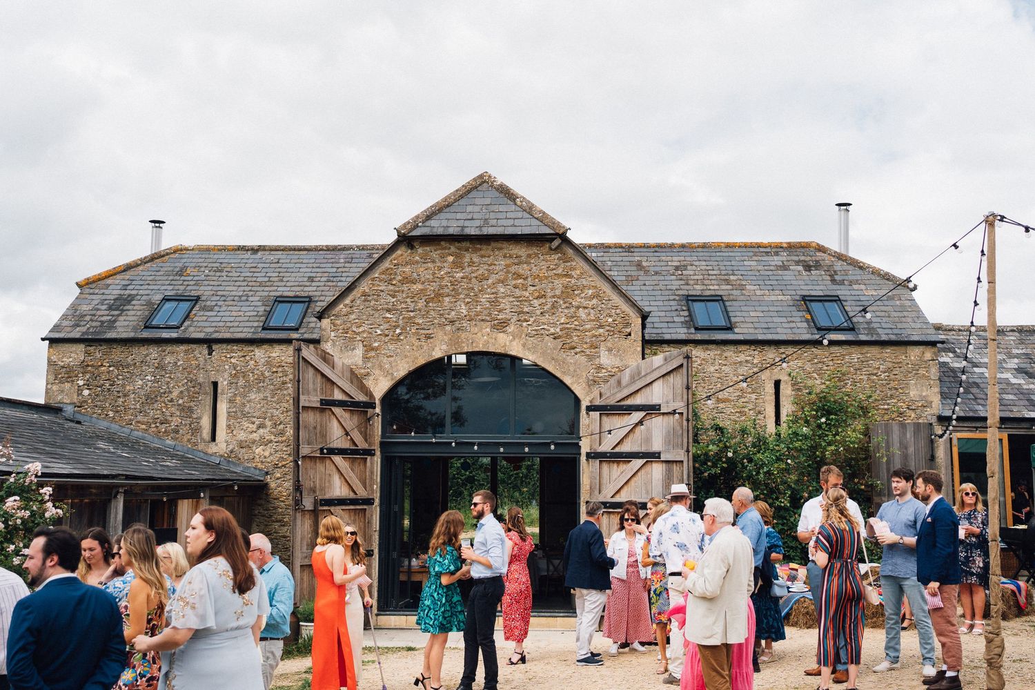Guests mingling outside a rustic stone barn wedding venue under cloudy skies with string lights and wooden doors.
