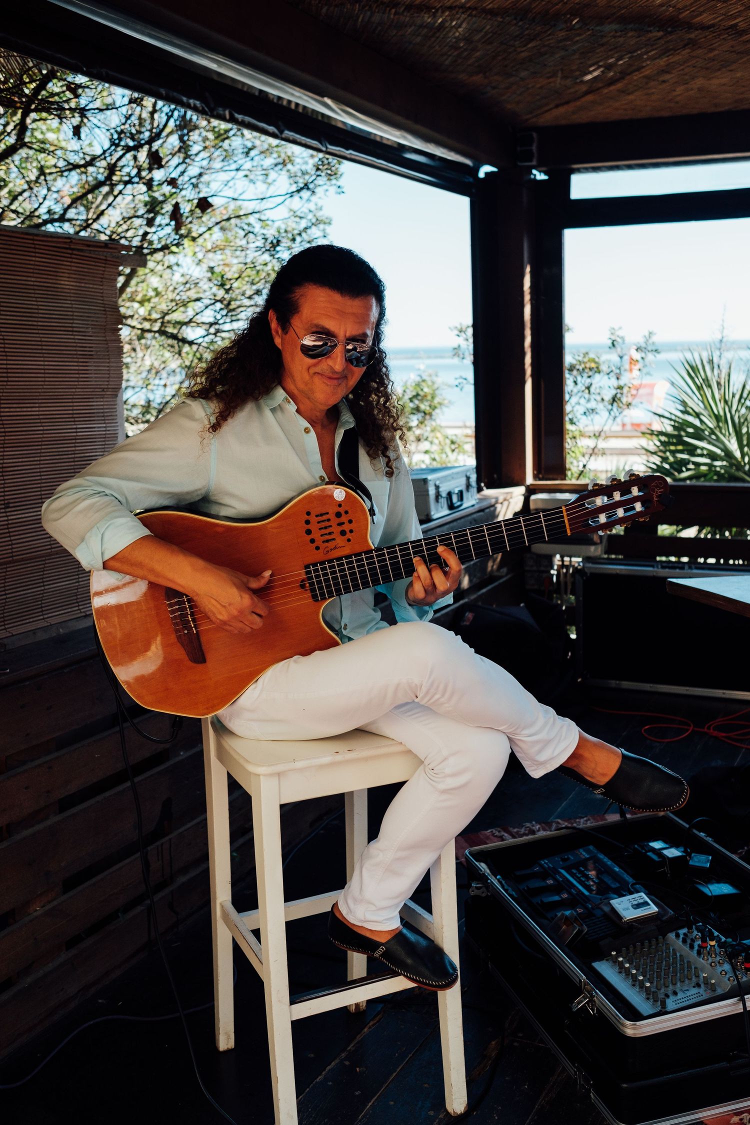 Musician in white clothing playing acoustic guitar while seated indoors near a window.