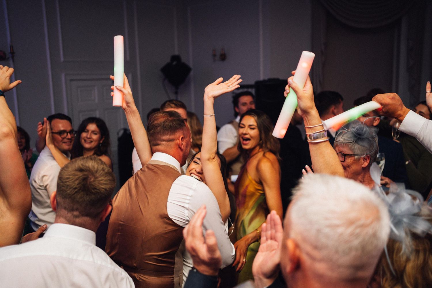 Energetic wedding party dancing with hands in the air under purple and blue lighting.